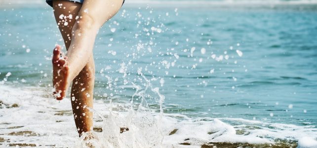Eine Frau läuft barfuss am Strand und spritzt mit den Füßen Wasser hoch